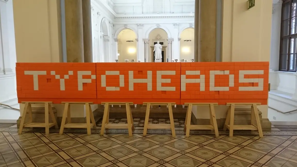 Schriftzug TYPOHEADS aus orangen und weißen Packungen Manner-Schnitten auf Holzböcken in einer Säulenhalle